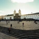 People crossing a broad square before a white church in Quito